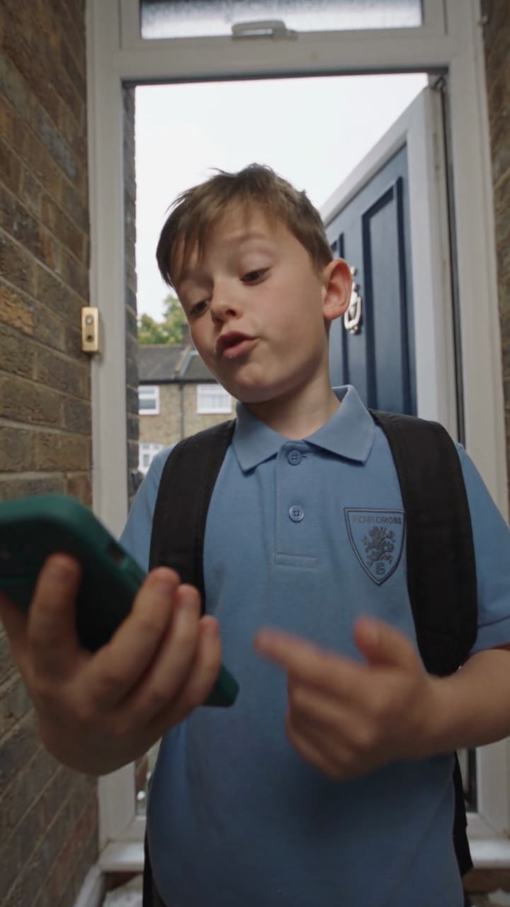 A boy in a school shirt looks at his phone in the front door.